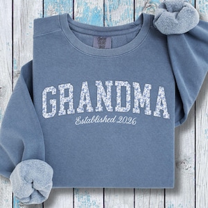 May include: A blue jean colored crewneck sweatshirt featuring the word GRANDMA in a white floral pattern across the chest, with the text Established 2026 printed in white underneath. The garment has long sleeves with ribbed cuffs and a crew neckline.