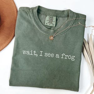 May include: A moss green t-shirt with the phrase "wait, I see a frog" printed in white. A gold necklace with a pendant rests on the shirt. A brown hat and dried leaves are also in the image.