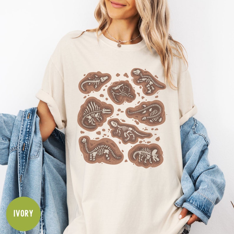 May include: A beige t-shirt featuring a graphic print of dinosaur skeletons in brown and white. The skeletons are in various poses and are surrounded by brown dirt. The word "IVORY" is written in green on a white circle in the bottom left corner of the image.