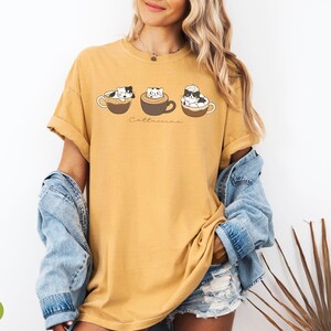 Puede incluir: Una camiseta amarilla mostaza con un gr&aacute;fico de tres gatos en tazas de caf&eacute;. El texto "Cat-uccino" est&aacute; impreso debajo de los gatos.