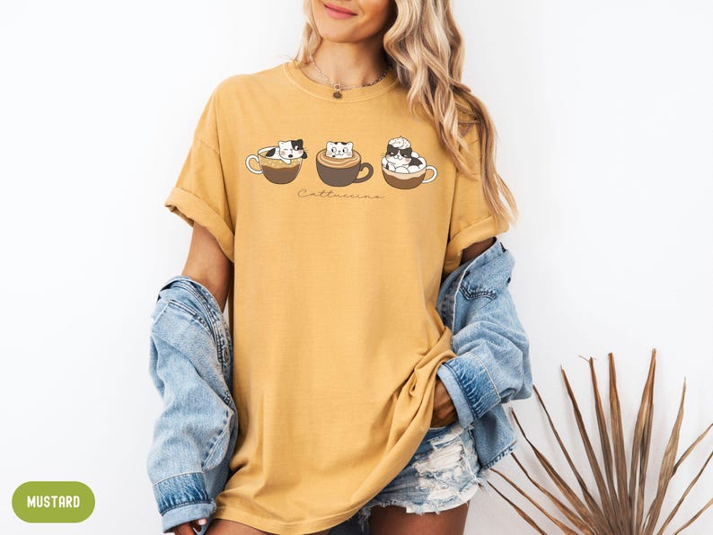 Puede incluir: Una camiseta amarilla mostaza con un gr&aacute;fico de tres gatos en tazas de caf&eacute;. El texto "Cat-uccino" est&aacute; impreso debajo de los gatos.