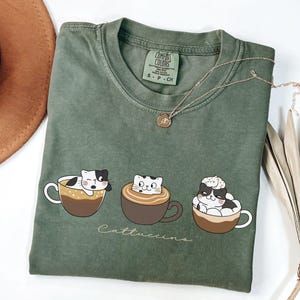 Puede incluir: Una camiseta verde con un gr&aacute;fico de tres gatos en tazas de caf&eacute;. El texto "Cattuccinos" est&aacute; impreso debajo de los gatos. La camiseta est&aacute; hecha por Comfort Colors y es talla S-P-CH.