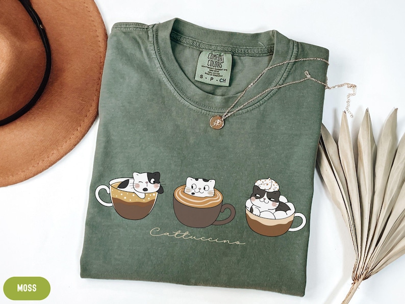 Puede incluir: Una camiseta verde con un gr&aacute;fico de tres gatos en tazas de caf&eacute;. El texto "Cattuccinos" est&aacute; impreso debajo de los gatos. La camiseta est&aacute; hecha por Comfort Colors y es talla S-P-CH.