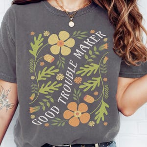 May include: A dark gray t-shirt with a floral design and the text "Good Trouble Maker" in white.