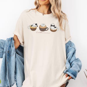 Puede incluir: Una camiseta blanca con un gr&aacute;fico de tres gatos en tazas de caf&eacute;. El texto "Catteccino" est&aacute; impreso debajo de los gatos. La camiseta est&aacute; siendo modelada por una persona que lleva una chaqueta vaquera.
