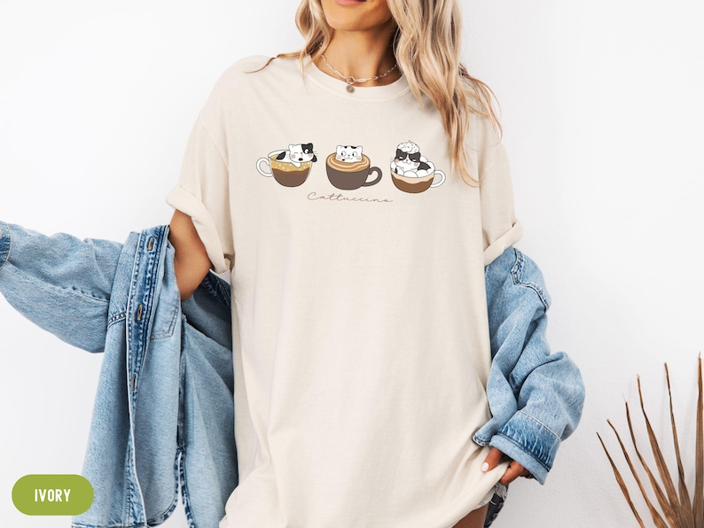 Puede incluir: Una camiseta blanca con un gr&aacute;fico de tres gatos en tazas de caf&eacute;. El texto "Catteccino" est&aacute; impreso debajo de los gatos. La camiseta est&aacute; siendo modelada por una persona que lleva una chaqueta vaquera.