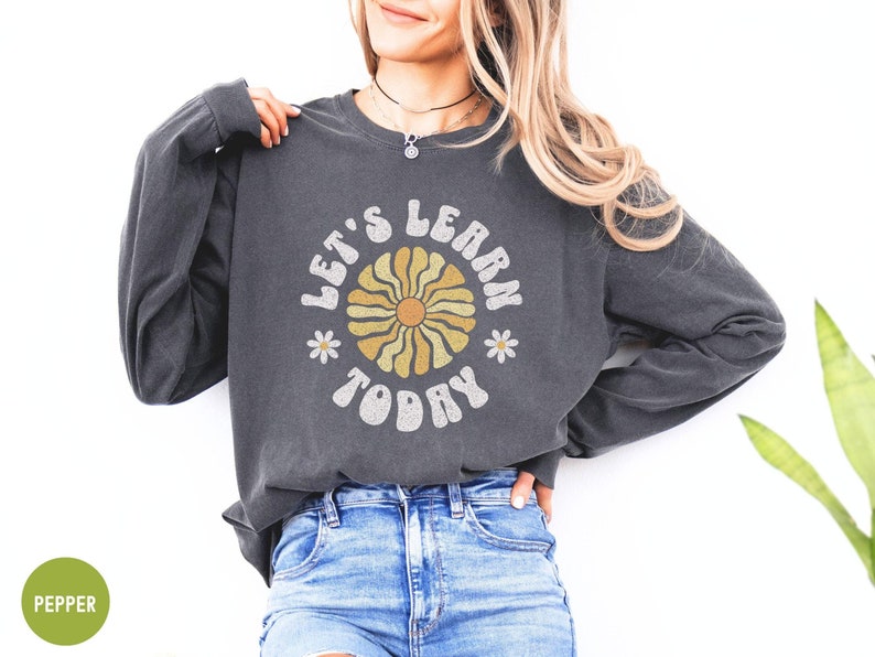May include: Dark gray long-sleeve sweatshirt with the text "LET'S LEARN TODAY" in a retro font. The design includes a yellow and tan flower with daisy accents. The sweatshirt is a comfortable, casual style.