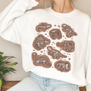 May include: White sweatshirt with a design of brown and white dinosaur skeletons. The graphic shows various dinosaur fossils in a dirt-like setting. The sweatshirt is a long-sleeved, crew neck style.