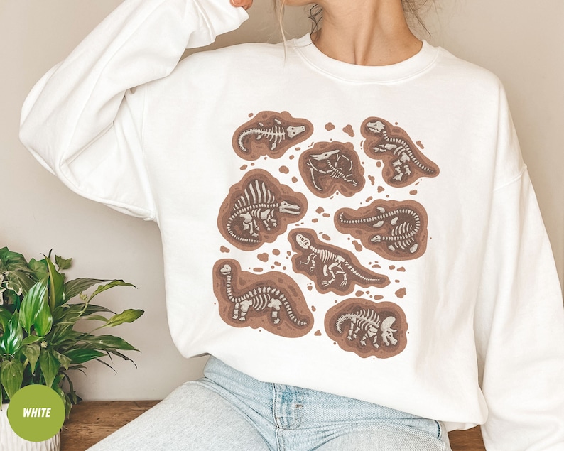 May include: White sweatshirt with a design of brown and white dinosaur skeletons. The graphic shows various dinosaur fossils in a dirt-like setting. The sweatshirt is a long-sleeved, crew neck style.