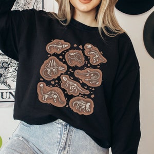 May include: Black sweatshirt with a design of dinosaur fossils in a brown, earthy pattern. The fossils are illustrated in detail, showing each dinosaur skeleton. The sweatshirt is a casual garment, suitable for everyday wear. The design is in a scientific style.