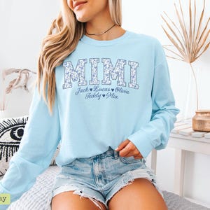 May include: Pale blue long-sleeved sweatshirt featuring the word "MIMI" in a floral pattern. Underneath, the names Jack, Lucas, Olivia, Teddy, and Mia are displayed. The sweatshirt is worn with denim shorts.