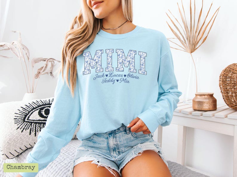May include: Pale blue long-sleeved sweatshirt featuring the word "MIMI" in a floral pattern. Underneath, the names Jack, Lucas, Olivia, Teddy, and Mia are displayed. The sweatshirt is worn with denim shorts.