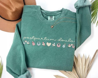 May include: A sage green crewneck sweatshirt featuring the text "postpartum doula" in a light cream script font, accompanied by a row of small, colourful icons including a stork, baby bottle, bib, pacifier, heart, and rattle.