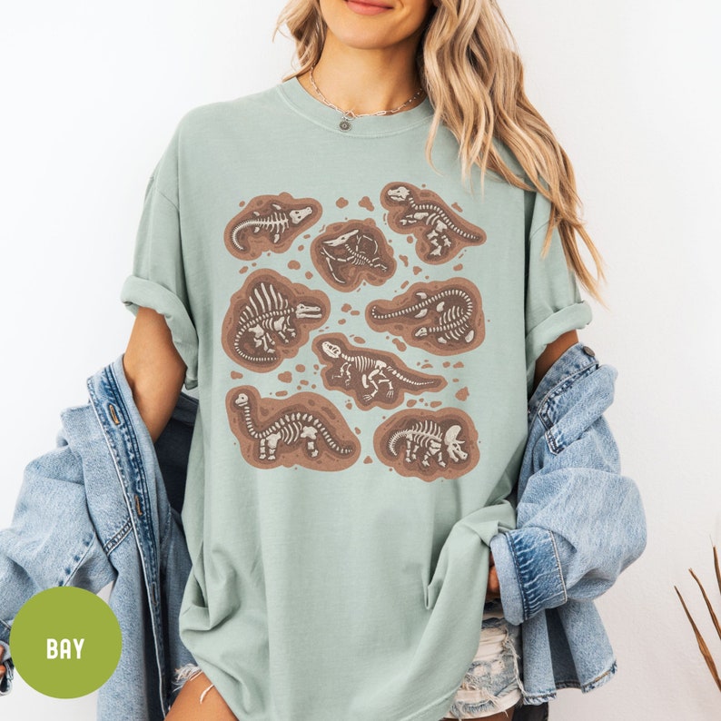 May include: A light green t-shirt featuring a graphic print of dinosaur skeletons in various poses. The skeletons are brown and white and are set against a light brown background. The shirt has a crew neck and short sleeves.