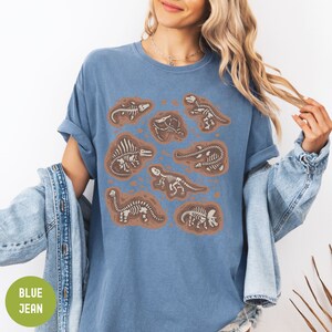 May include: A blue jean t-shirt featuring a print of various dinosaur skeletons in brown and white. The skeletons are depicted in different poses and sizes, outlined in brown.