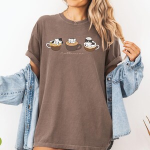 Puede incluir: Una camiseta marr&oacute;n con un gr&aacute;fico de tres gatos en tazas de caf&eacute;. El texto "Catteccino" est&aacute; impreso debajo de los gatos.