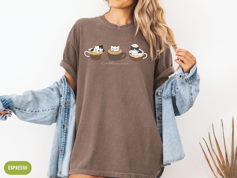 Puede incluir: Una camiseta marr&oacute;n con un gr&aacute;fico de tres gatos en tazas de caf&eacute;. El texto "Catteccino" est&aacute; impreso debajo de los gatos.