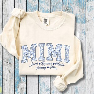 May include: Cream-coloured long-sleeved sweatshirt featuring the word "MIMI" in blue block letters filled with a butterfly pattern. Underneath are the names Jack, Lucas, Olivia, Teddy, and Mia.