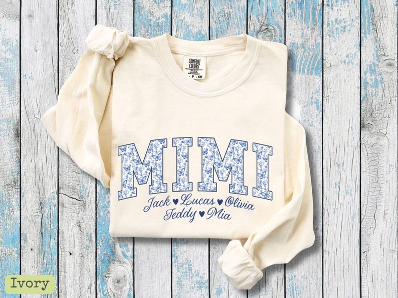 May include: Cream-coloured long-sleeved sweatshirt featuring the word "MIMI" in blue block letters filled with a butterfly pattern. Underneath are the names Jack, Lucas, Olivia, Teddy, and Mia.