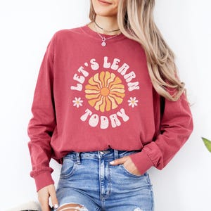 May include: Crimson long-sleeve sweatshirt with the text "LET'S LEARN TODAY" in a retro font. The design includes a yellow and orange sun graphic and small white flowers. The sweatshirt is paired with blue jeans.