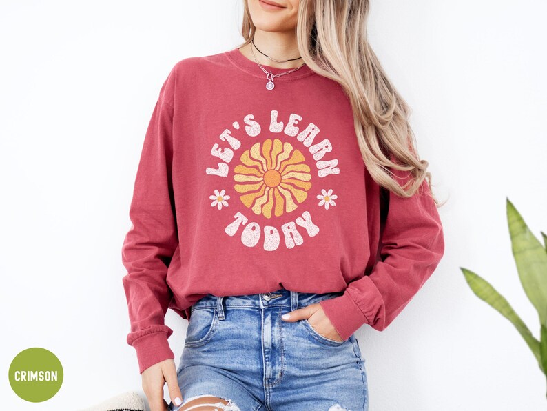 May include: Crimson long-sleeve sweatshirt with the text "LET'S LEARN TODAY" in a retro font. The design includes a yellow and orange sun graphic and small white flowers. The sweatshirt is paired with blue jeans.