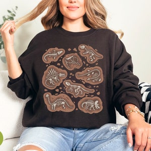 May include: A dark brown sweatshirt with a design of dinosaur fossils in a brown, earthy style. The fossils are white, contrasting with the dark background. The sweatshirt has a crew neck and long sleeves.