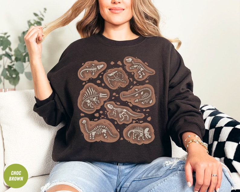 May include: A dark brown sweatshirt with a design of dinosaur fossils in a brown, earthy style. The fossils are white, contrasting with the dark background. The sweatshirt has a crew neck and long sleeves.