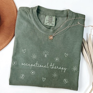 May include: A green t-shirt with the text "occupational therapy" in white script. The shirt is decorated with small white drawings of hearts, flowers, butterflies, suns, and light bulbs.