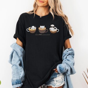 Puede incluir: Camiseta negra con un gr&aacute;fico de tres gatos en tazas de caf&eacute;. El texto "Cattuccino" est&aacute; impreso debajo de las tazas.