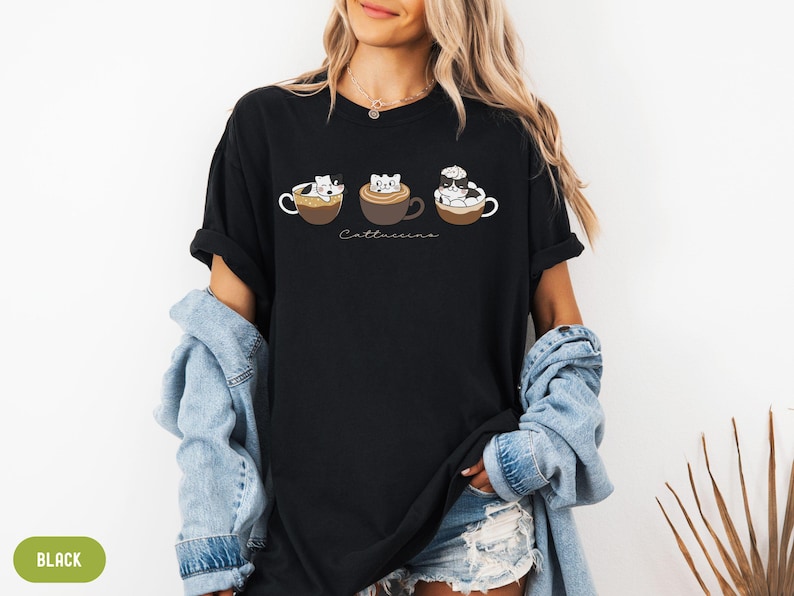 Puede incluir: Camiseta negra con un gr&aacute;fico de tres gatos en tazas de caf&eacute;. El texto "Cattuccino" est&aacute; impreso debajo de las tazas.