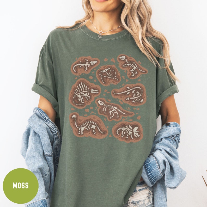 May include: A moss green t-shirt featuring a graphic print of dinosaur skeletons in various poses. The skeletons are outlined in white and are set against a brown background.