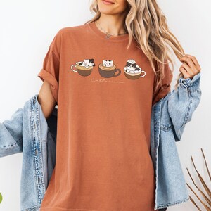 Puede incluir: Una camiseta de color &oacute;xido con un gr&aacute;fico de tres gatos en tazas de caf&eacute;. El texto "Cat-uccino" est&aacute; impreso debajo del gr&aacute;fico.