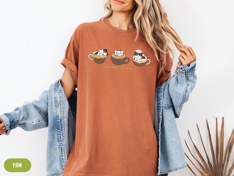 Puede incluir: Una camiseta de color &oacute;xido con un gr&aacute;fico de tres gatos en tazas de caf&eacute;. El texto "Cat-uccino" est&aacute; impreso debajo del gr&aacute;fico.