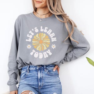 May include: A grey long-sleeve shirt with the text "LET'S LEARN TODAY" in a retro font, featuring a flower design. The shirt is paired with blue jeans. The overall style is casual and comfortable.