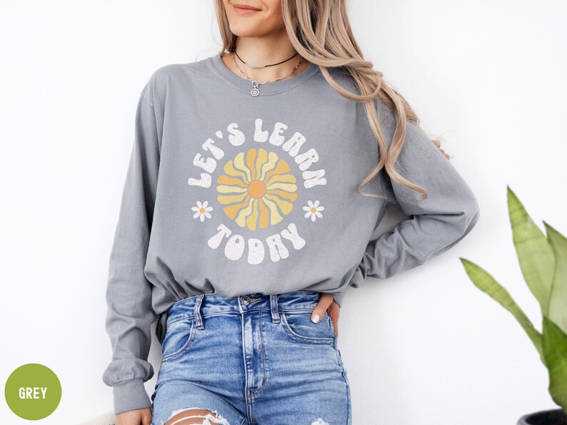 May include: A grey long-sleeve shirt with the text "LET'S LEARN TODAY" in a retro font, featuring a flower design. The shirt is paired with blue jeans. The overall style is casual and comfortable.