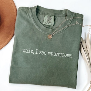 May include: A moss green t-shirt with the phrase "wait, I see mushrooms" in white. A gold necklace with a pendant and a brown hat are next to the shirt. A dried palm leaf is also in the image.