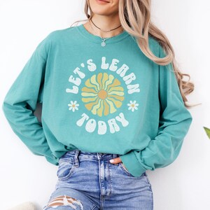 May include: A teal long-sleeve shirt with the words "LET'S LEARN TODAY" in white, arched around a retro flower design in yellow and orange. The shirt is paired with blue jeans. The overall style is casual and comfortable.