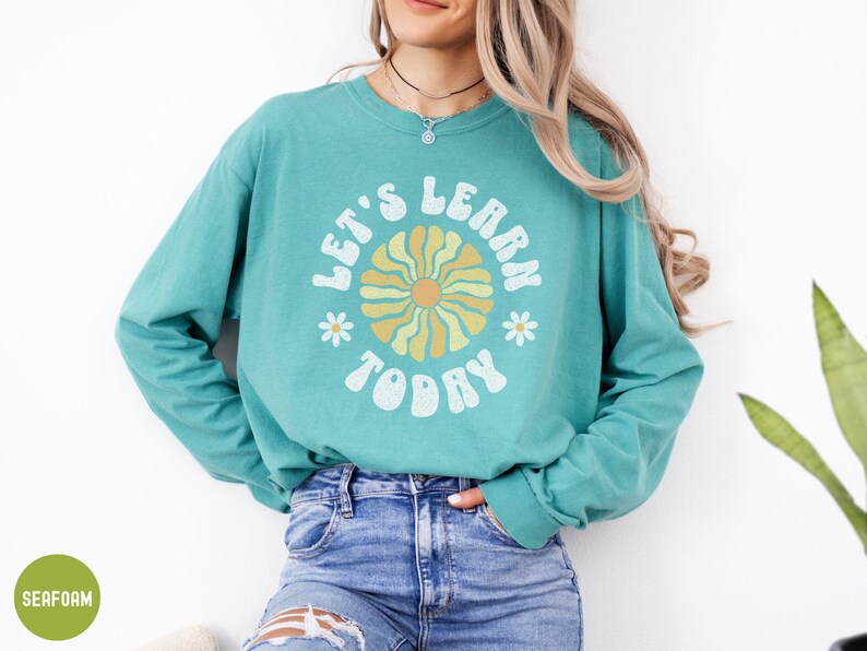May include: A teal long-sleeve shirt with the words "LET'S LEARN TODAY" in white, arched around a retro flower design in yellow and orange. The shirt is paired with blue jeans. The overall style is casual and comfortable.