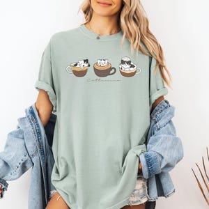 Puede incluir: Una camiseta verde claro con un gr&aacute;fico de tres gatos en tazas de caf&eacute;. El texto "Cat-t-uccino" est&aacute; impreso debajo de los gatos.