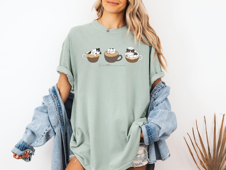 Puede incluir: Una camiseta verde claro con un gr&aacute;fico de tres gatos en tazas de caf&eacute;. El texto "Cat-t-uccino" est&aacute; impreso debajo de los gatos.