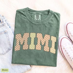 May include: A moss green crew neck t-shirt featuring the word MIMI in a large, arched varsity font. The letters have a tan and white gingham check pattern with a thin dark outline. The shirt has a Comfort Colors brand label at the neck.