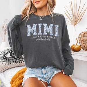 May include: Charcoal grey long-sleeved sweatshirt featuring the word "MIMI" in a floral pattern. Underneath, the names Jack, Lucas, Olivia, Teddy, and Mia are printed. A relaxed, casual style of top.