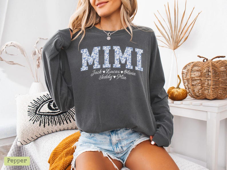 May include: Charcoal grey long-sleeved sweatshirt featuring the word "MIMI" in a floral pattern. Underneath, the names Jack, Lucas, Olivia, Teddy, and Mia are printed. A relaxed, casual style of top.