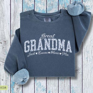 Pu&ograve; includere: Una felpa blu denim con la scritta "Great Grandma" in bianco, con lettere a fantasia floreale. Sotto ci sono i nomi "Jack", "Lucas", "Olivia" e "Mia". La felpa ha un girocollo e maniche lunghe.