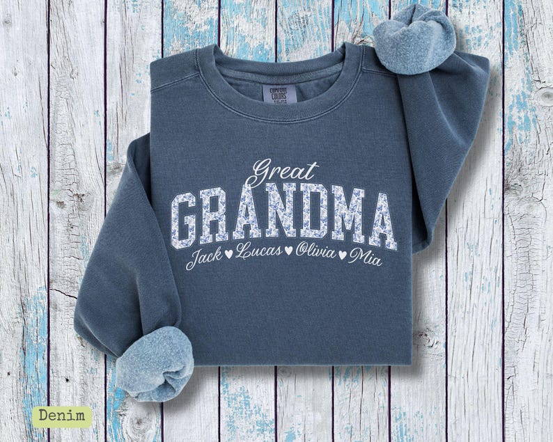 Pu&ograve; includere: Una felpa blu denim con la scritta "Great Grandma" in bianco, con lettere a fantasia floreale. Sotto ci sono i nomi "Jack", "Lucas", "Olivia" e "Mia". La felpa ha un girocollo e maniche lunghe.