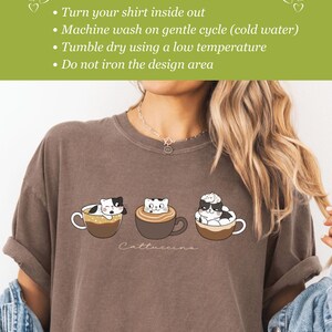 Puede incluir: Una camiseta marr&oacute;n con un gr&aacute;fico de tres gatos en tazas de caf&eacute;. El texto "Cattuccina" est&aacute; impreso debajo del gr&aacute;fico. Las instrucciones de cuidado est&aacute;n impresas en un fondo verde en la parte superior de la imagen. Las instrucciones son: Dale la vuelta a tu camiseta, Lava a m&aacute;quina en ciclo suave (agua fr&iacute;a), Seca en secadora a baja temperatura, No planches el &aacute;rea del dise&ntilde;o.