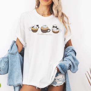 Puede incluir: Camiseta blanca con un gr&aacute;fico de tres gatos en tazas de caf&eacute;. El texto "Catteccino" est&aacute; impreso debajo del gr&aacute;fico.