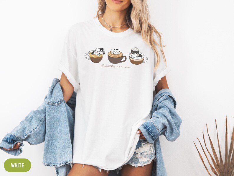 Puede incluir: Camiseta blanca con un gr&aacute;fico de tres gatos en tazas de caf&eacute;. El texto "Catteccino" est&aacute; impreso debajo del gr&aacute;fico.