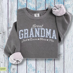 Pu&ograve; includere: Una felpa grigia con le parole "Great Grandma" in corsivo bianco e stampa floreale blu. I nomi Jack, Lucas, Olivia e Mia sono stampati sotto la parola Grandma. Le maniche sono arrotolate.