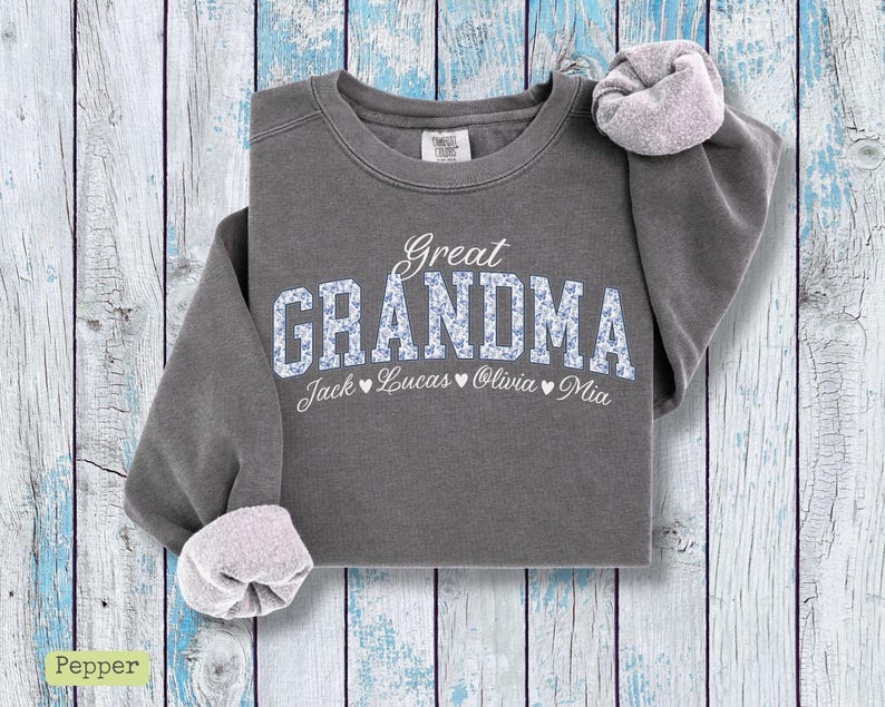 Pu&ograve; includere: Una felpa grigia con le parole "Great Grandma" in corsivo bianco e stampa floreale blu. I nomi Jack, Lucas, Olivia e Mia sono stampati sotto la parola Grandma. Le maniche sono arrotolate.